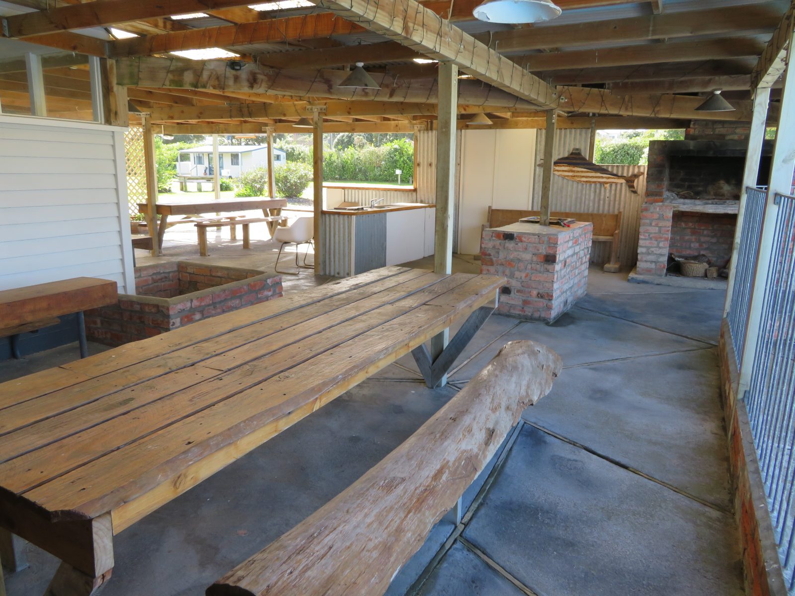 Covered outdoor kitchen area with long wooden tables, bench seating and a brick barbecue fireplace